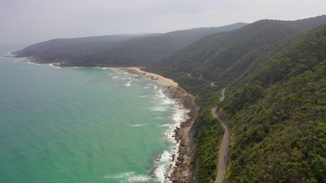 在阳光明媚的日子里，航拍海上绿色山脉上的道路，无人机向后飞行，对着天空的海岸线-大洋路，澳大利亚视频素材