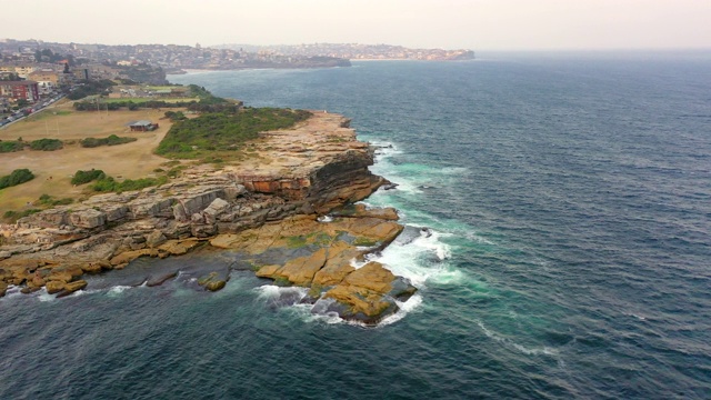 空中平移拍摄的海浪拍打岩石的建筑对天空，无人机飞过附近的城市-悉尼，澳大利亚视频素材