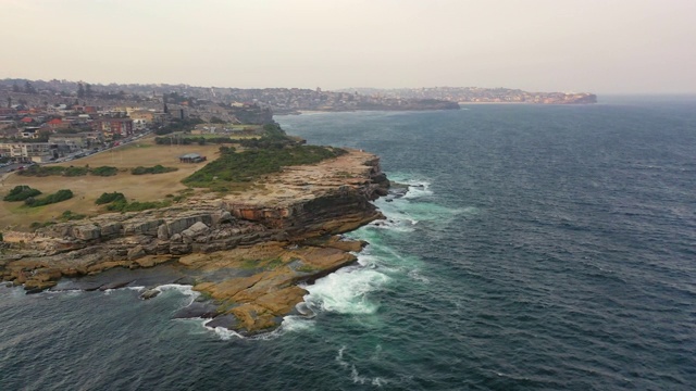 空中平移拍摄的岩石在海洋和城市对天空，无人机飞过海浪溅在海岸线上-悉尼，澳大利亚视频素材