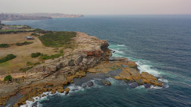 空中摇摄海浪拍打岩石附近的城市对天空，无人机飞越海洋-悉尼，澳大利亚视频素材