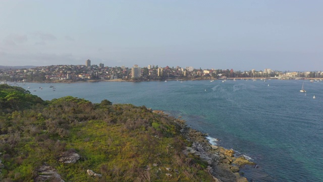 空中向前拍摄的蓝色海洋上的船只由建筑物对天空，无人机飞过植物附近的城市-悉尼，澳大利亚视频素材