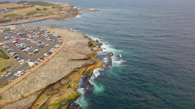 空中倾斜拍摄人们在海上和停车场的岩石上，无人机在溅起的海浪中向前飞行——澳大利亚悉尼视频素材