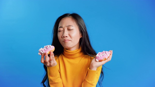 饥饿漂亮的亚洲女孩吃油炸圈饼在蓝色的背景视频素材