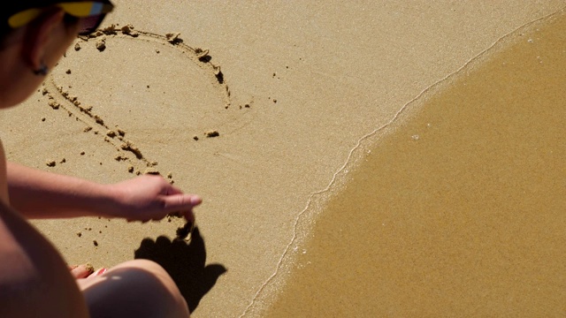 一个年轻女子在沙滩上画了一颗心。海浪来了视频素材