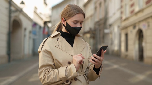 年轻的金发女人在防护医疗面具走在空的街道上。用手机发短信，滚轴上网。搜索新冠肺炎疫情防控城市。视频素材