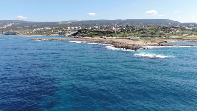 广阔的镜头塞浦路斯旅游胜地在地中海海岸。鸟瞰图上的小建筑散布在绿色的山丘上，海岸线布满了沙子和岩石。夏日阳光下湛蓝的海水。视频素材