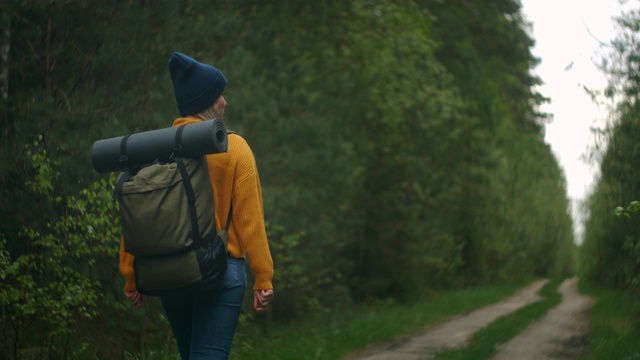 慢镜头:一名年轻女子在秋天的森林里徒步旅行。积极健康的白种女性背着背包，拿着木头。女性旅行者与徒步森林后视图，休闲，生物旅游，徒步旅行视频素材