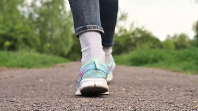 穿着运动鞋的女人走在一条土路上。穿着牛仔裤和运动鞋的女孩正在穿过公园。特写腿，一个女孩在走路视频素材