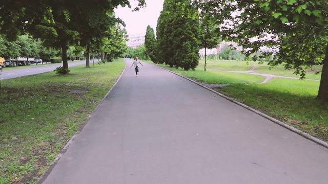 扎着马尾辫的女孩在镜头前穿过公园的马路。夏天快乐微笑的孩子。手持式稳定凸轮飞凸轮运动回拍摄视频素材