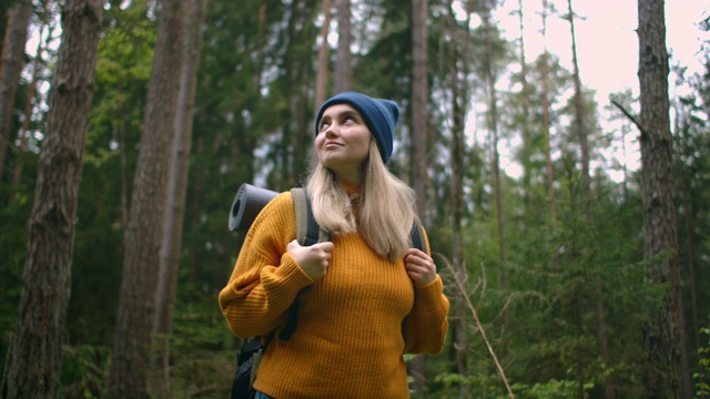 一位穿着黄色毛衣、背着背包的女性旅行者正走在森林里的一条小路上，欣赏着美丽的景色。女性旅行者探索森林在毛衣和背包视频素材