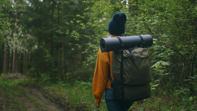 一位穿着黄色毛衣、背着背包的女性旅行者正走在森林里的一条小路上，欣赏着美丽的景色。女性旅行者探索森林在毛衣和背包视频素材