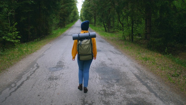 一位穿着黄色毛衣、背着背包的女性旅行者正走在森林里的一条小路上，欣赏着美丽的景色。女性旅行者探索森林在毛衣和背包视频素材