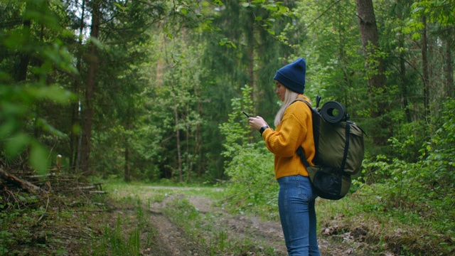 慢镜头:一名年轻女子在森林里用智能手机享受周末。旅行的女人背着背包走在森林的小路上，在绿色的树林里看电话和地图视频素材