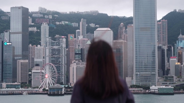 后视图的年轻亚洲女子站在维多利亚港的海滨长廊，看香港城市的天际线。将焦点从前景逐渐转移到背景视频素材