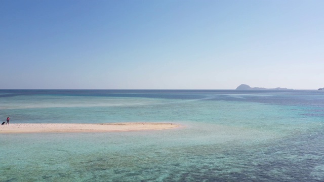 宽镜头女子走在拉着行李沿着空旷的岛屿海滩被海洋包围视频素材