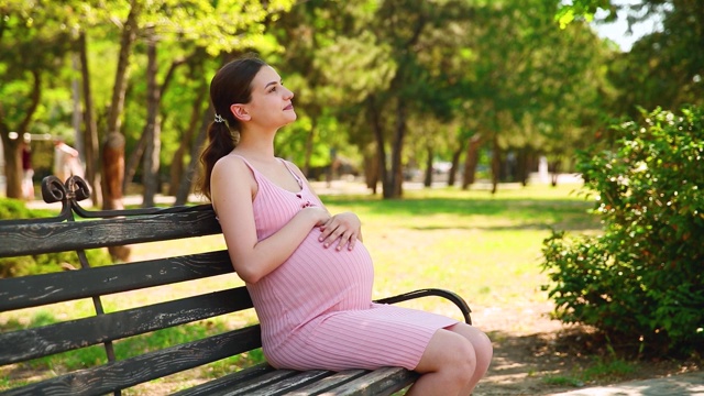 一位孕妇在绿色的公园里抚摸着自己的肚子。一个快乐的怀孕女孩坐在公园的长椅上，用他的手轻轻地抚摸着他的肚子，这是一个夏日阳光明媚的日子视频素材