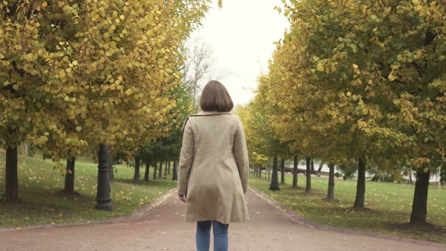 穿着大衣的女人在秋天的公园里沿着树走视频素材