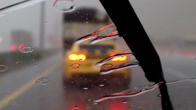 雨落在汽车挡风玻璃上。在暴雨暴雨中驾驶汽车行驶在交通堵塞的街道上，车内一辆汽车行驶着，模糊了交通灯的背景。视频素材