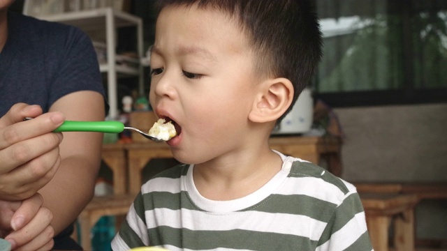 亚洲男孩看起来很可爱，坐在那里吃早餐，他的妈妈在喂他。视频素材