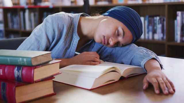 亚洲女学生戴着蓝色的头巾坐在书桌前，翻开书睡觉视频素材