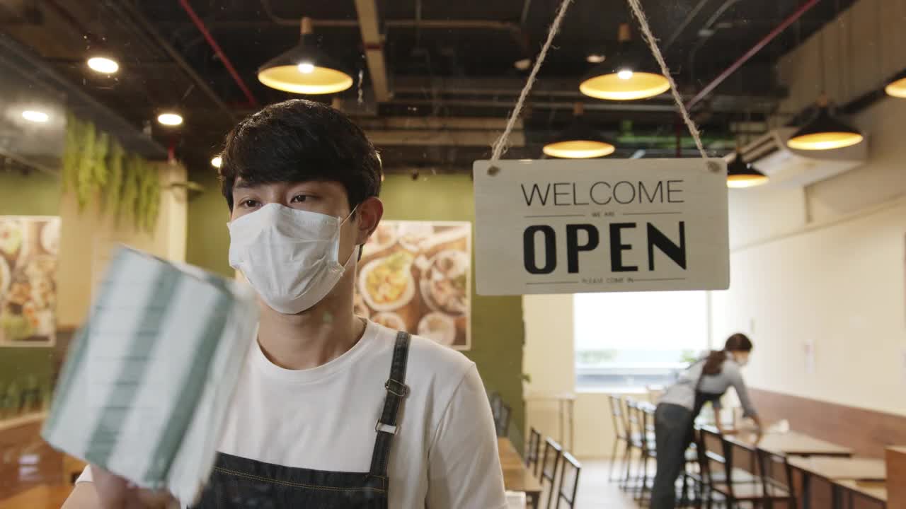 在封锁和冠状病毒covid-19疫情后，一名亚洲男子在一家餐馆工作，打开前面的开放标志开门视频购买