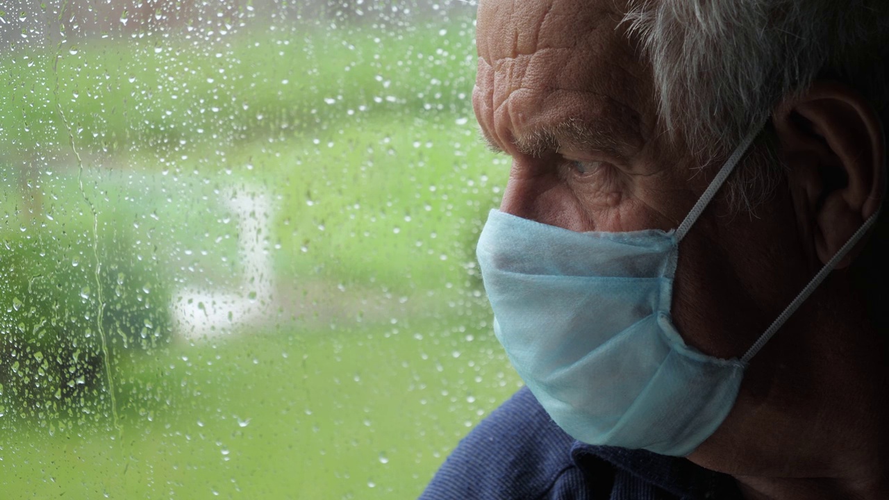 下雨时，戴着医用防护口罩的悲伤老人望着窗外的街道视频下载