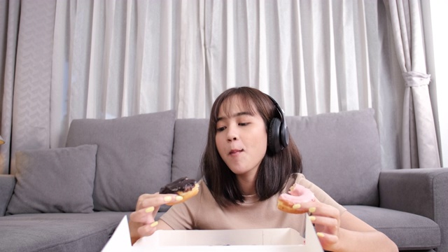 年轻的亚洲女性在沙发上吃甜甜圈垃圾食品，在客厅听音乐，快餐外卖服务安全，通过社交距离概念阻止冠状病毒的传播。视频素材