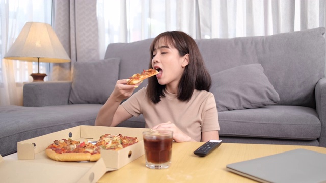 年轻的亚洲女性外卖吃垃圾食品比萨饼与饮料可乐和使用遥控器，女性享受快乐和看电视在家，快餐配送服务理念。视频素材