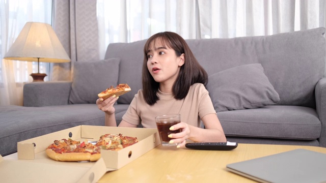 年轻的亚洲女人外卖垃圾食品吃披萨和饮料可乐。女性享受在家里吃和看电视的乐趣。快餐配送服务理念。视频素材