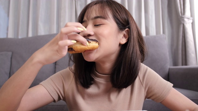 年轻的亚洲女性在客厅外卖吃甜甜圈垃圾食品在家享受幸福，快餐外卖服务安全，通过社交距离概念阻止冠状病毒的传播。视频素材