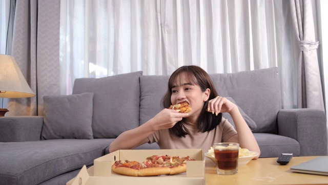 年轻的亚洲女人外卖垃圾食品吃披萨和饮料可乐，女性享受幸福和看电视在家。快餐配送服务理念。视频素材