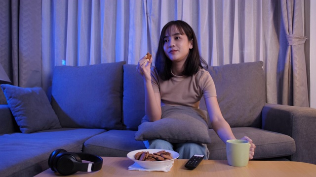 年轻的亚洲女性外卖吃垃圾食品，在沙发上看电视剧，吃快餐，在客厅喝酒，在家享受幸福。视频素材