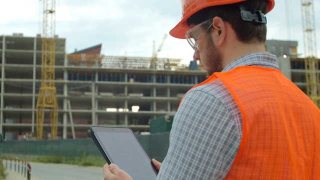 现代建筑工程师或建筑师戴着头盔在建筑工地使用工业电子平板工作。工程师的特写肖像与小工具的背景下的建筑。视频素材