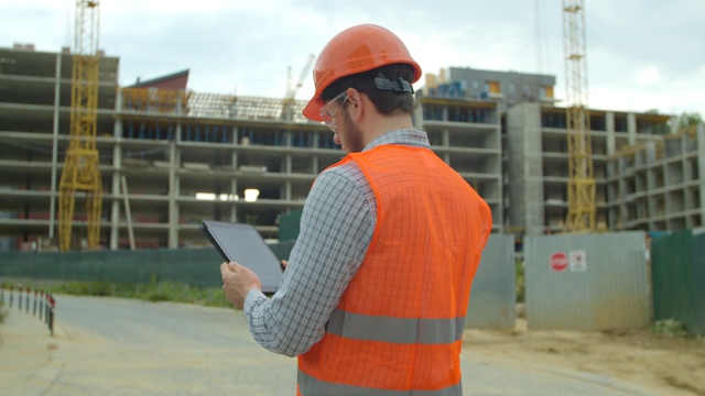 工程师的特写肖像与小工具的背景下的建筑。现代建筑工程师或建筑师戴着头盔在建筑工地使用工业电子平板工作。视频素材