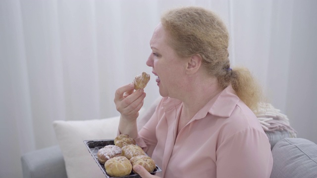 郁闷的胖女人嚼着甜面包哭。压力大的白人老年妇女有抑郁症的侧面。悲伤、垃圾食品、压力饮食视频素材