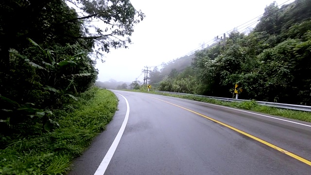 行驶在有黄色标记的柏油路上，穿过雨雾缭绕的热带雨林山脉。慢动作视频素材