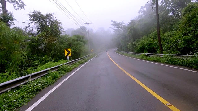 行驶在有黄色标记的柏油路上，穿过雨雾缭绕的热带雨林山脉。慢动作视频素材