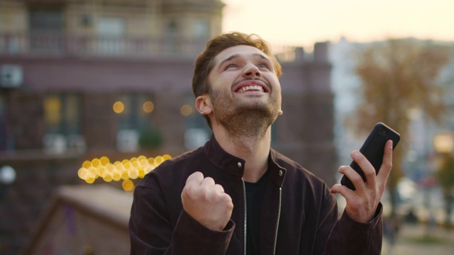 一个在户外接受短信电话的兴高采烈的家伙。激动的男人像胜利者一样手势视频素材