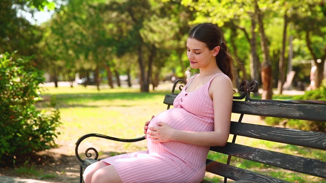 一位孕妇在绿色的公园里抚摸着自己的肚子。一个快乐的怀孕女孩坐在公园的长椅上，用他的手轻轻地抚摸着他的肚子，这是一个夏日阳光明媚的日子视频素材