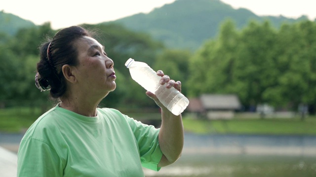 亚洲老年女性在公园运动后喝瓶装水，站在户外运动后放松，快乐活跃的退休理念，运动后提神，健康的生活方式视频素材