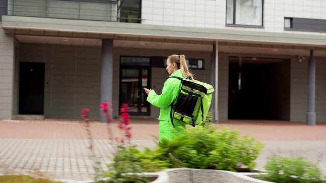 跟踪拍摄的女性青少年食品快递员身穿绿色制服，带着保暖袋，通过智能手机上的导航应用程序走在街上，按门铃进入大楼并送餐视频素材