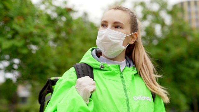 用慢镜头拍摄送餐女孩穿着绿色制服，戴着面罩和防护手套，背着保暖袋走在街上，回头看视频素材