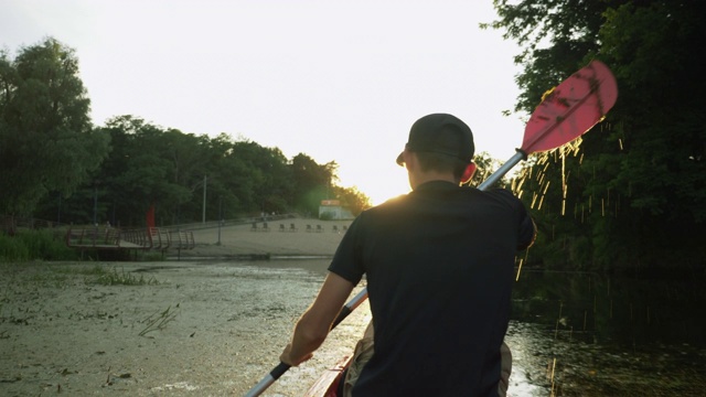 在金色夕阳的映衬下，一名男子正沿着静静的河流划着皮艇。男游客正在湖上划船。在极端活跃的假期期间，年轻的健康男子与桨漂浮在河上的独木舟视频素材