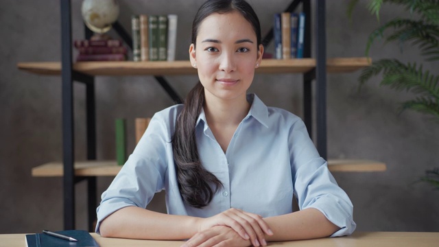 微笑的年轻亚洲女教师坐在桌子上，看着相机视频素材