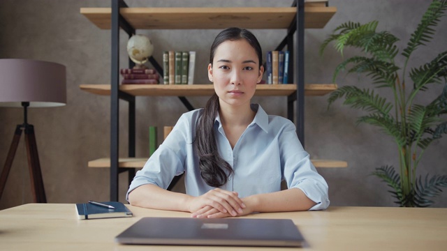 严肃的年轻亚洲女教师坐在桌子上，看着摄像机视频素材