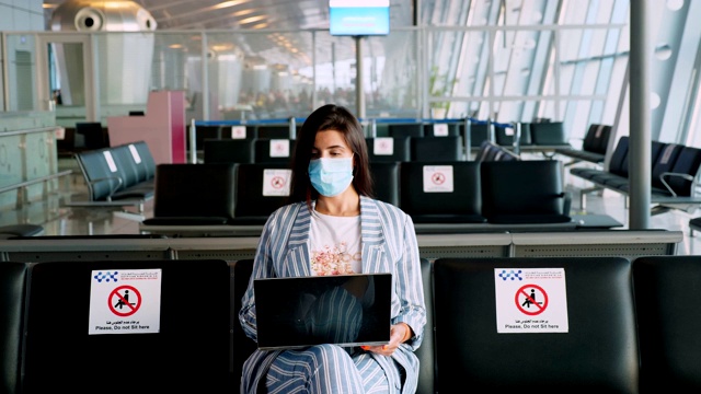 一名女性，戴着防护面具，坐在空空荡荡的机场休息室里等待登机，使用笔记本电脑。新冠肺炎疫情结束后，航空旅行重新开放。开放边界。旅游开放视频素材