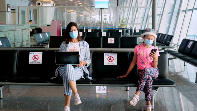 一名妇女和一名十几岁的女孩，戴着防护面具，坐在空空荡荡的机场休息室里等待登机，使用手机和笔记本电脑。新冠肺炎疫情结束后，航班重新开放。开放边界。旅游开放视频素材