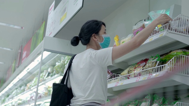 亚洲女性戴着防护口罩看货架上的食物、超市里的食品购物车。女顾客covid19新常态杂货店，从购物车看视频素材