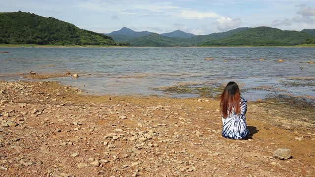 年轻的长发女子坐在岩石地上看风景，夏天蓝色的湖上的阿尔卑斯山，一缕头发移动，山脉，热带夏季生活。视频下载