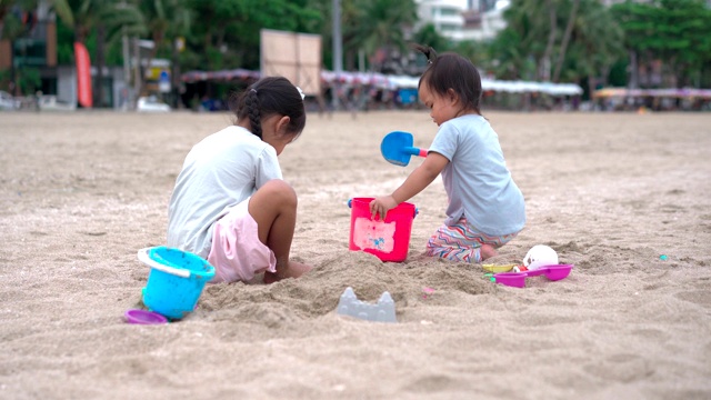 一个小女孩和她的小妹妹在沙滩上玩沙子，后面是城市的景色视频素材